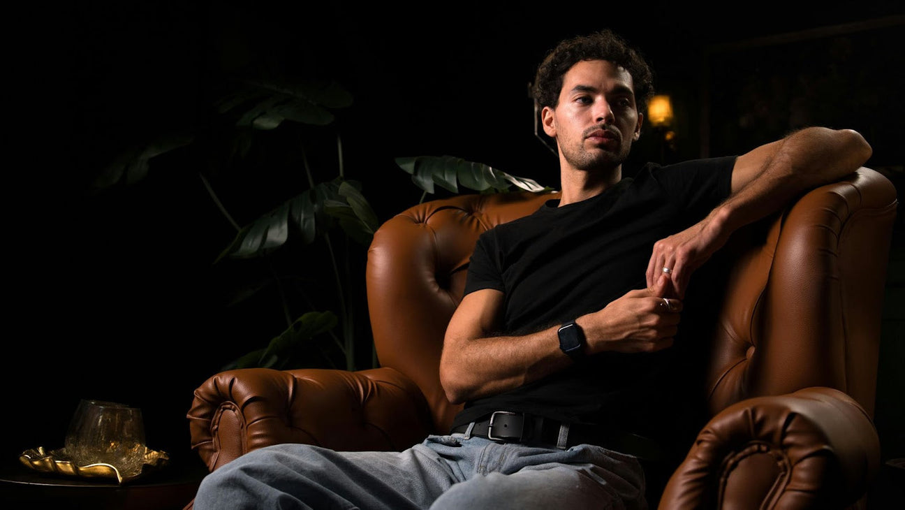 A stylish man showcasing modern streetwear fashion, wearing a black T-shirt, grey stacked joggers, and bold accessories, sitting on a brown leather chair.