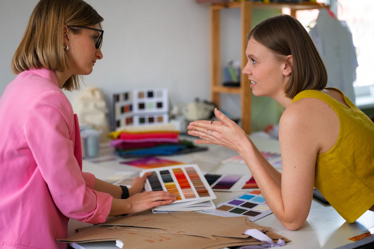 Two women discussing colour palettes at a fashion design studio, surrounded by fabric swatches and design materials.