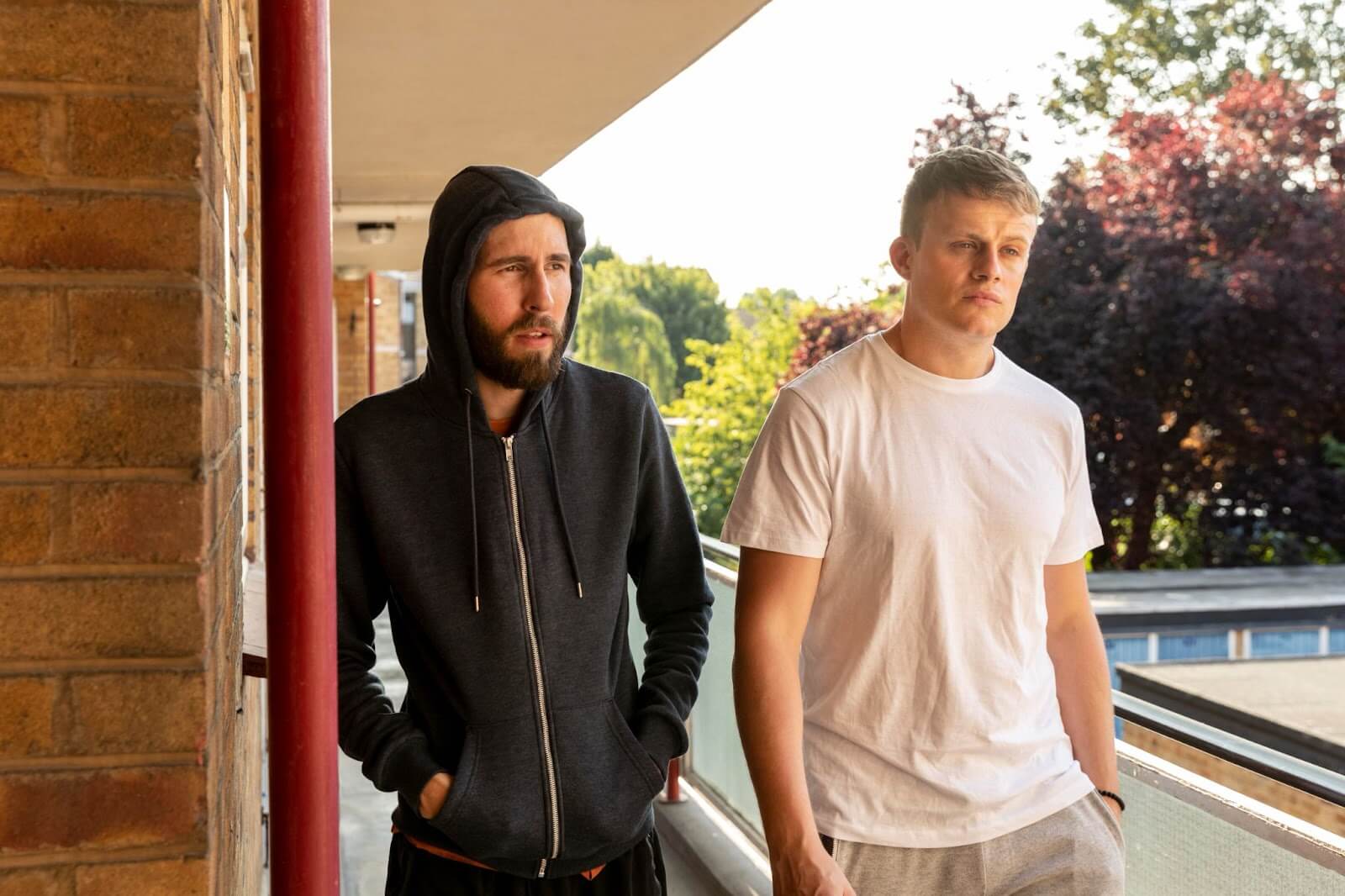 Two men standing on an outdoor walkway in a suburban area; one wears a black hoodie with the hood up, looking thoughtful, while the other wears a plain white T-shirt, looking contemplative.
