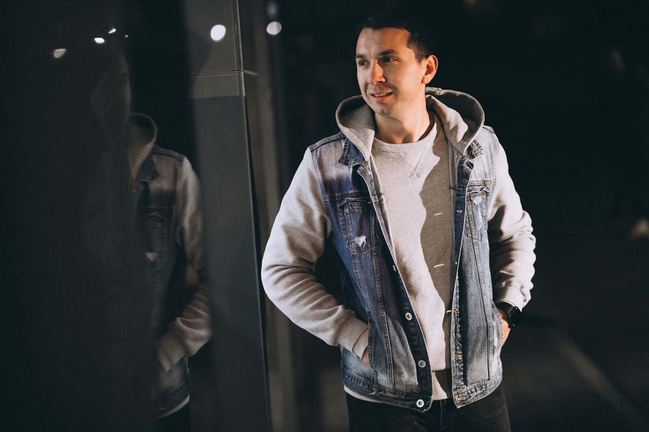 A young man wearing a casual outfit with a denim jacket layered over a hoodie, standing outdoors at night, smiling and looking away with his hands in his pockets. His reflection is visible in the glass wall beside him, creating a relaxed, urban vibe.