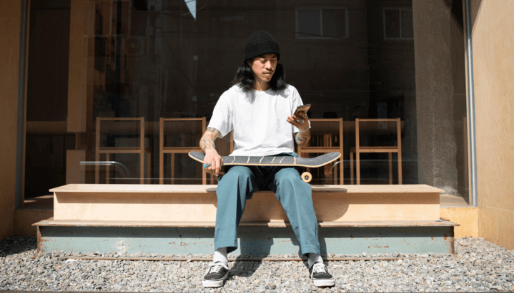 A young man with long hair and tattoos is sitting on steps, holding a skateboard on his lap, looking at his phone, and wearing a black beanie, white t-shirt, teal pants, and sneakers