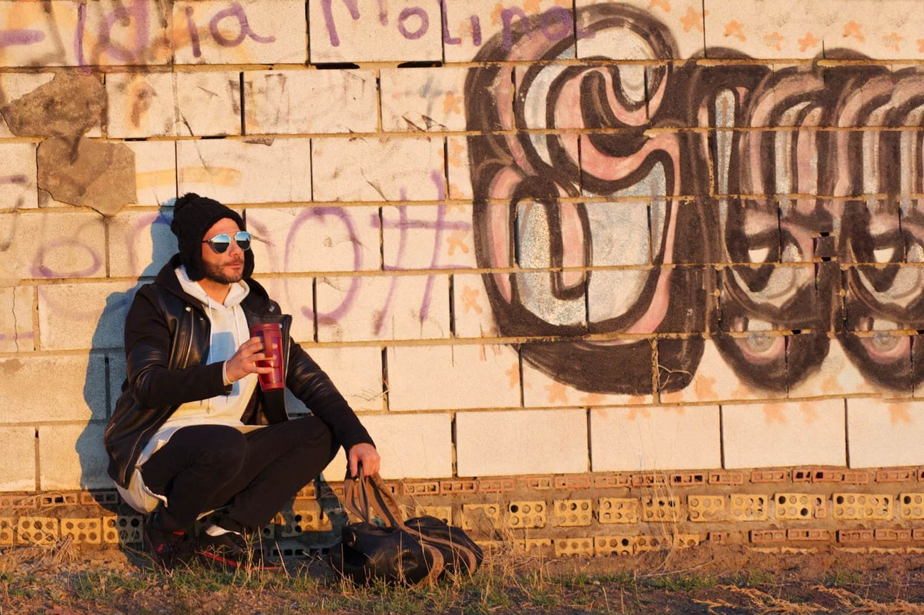 A stylish man dressed in casual streetwear, including a black leather jacket, hoodie, and beanie, sitting by a graffiti-covered wall, holding a thermos and carrying a duffle bag, reflecting the urban street style aesthetic.