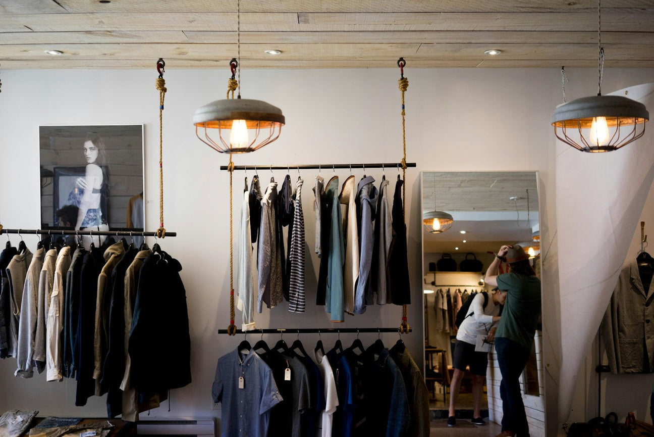 A modern streetwear clothing store with stylish apparel hanging on industrial-style racks, warm lighting, and a mirror reflecting shoppers.
