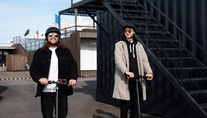 Two friends riding electric scooters, wearing casual streetwear with helmets on a sunny day.