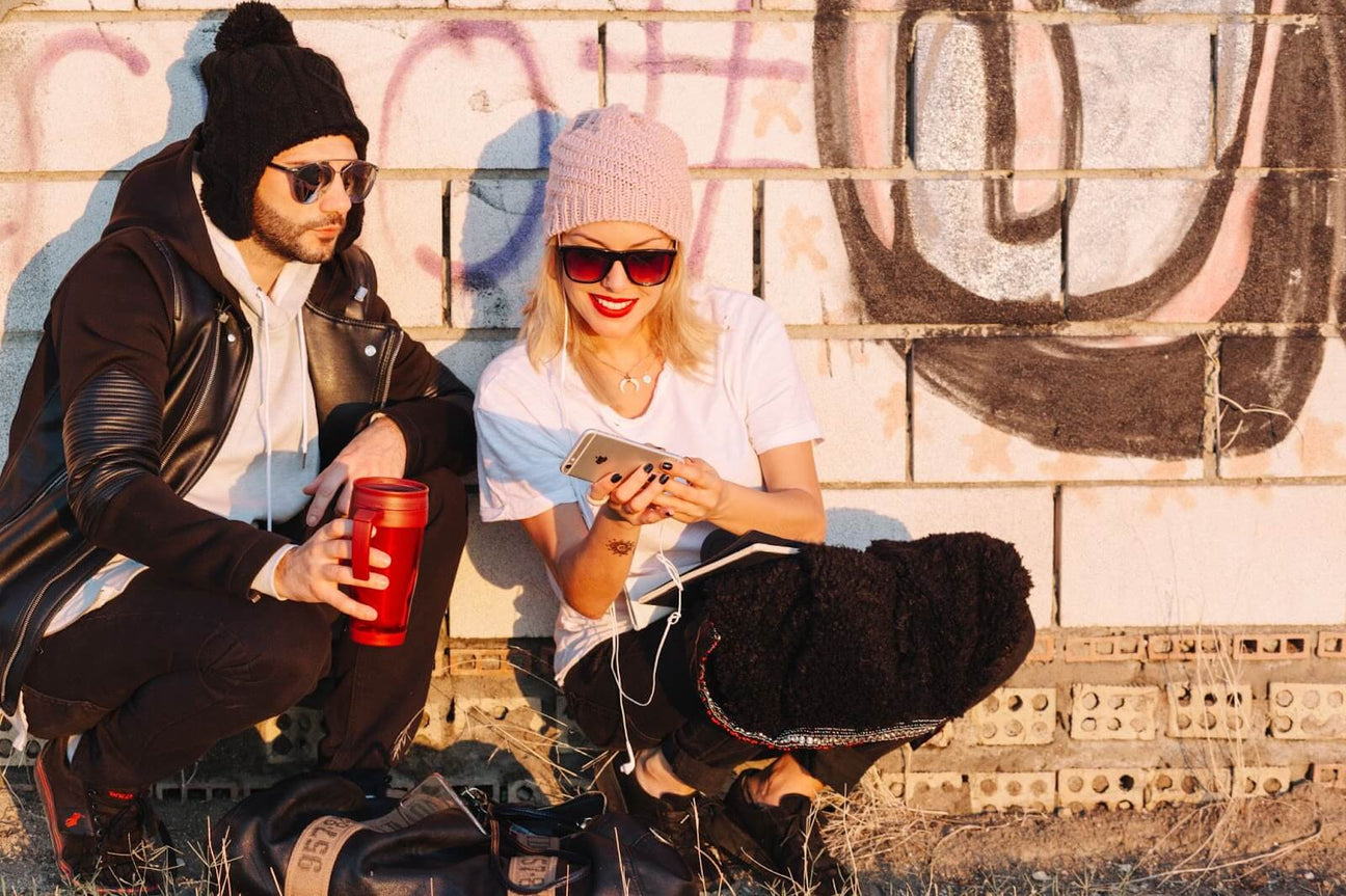 Young couple in streetwear sitting by a graffiti-covered brick wall, wearing beanies and sunglasses, enjoying a sunny moment.
