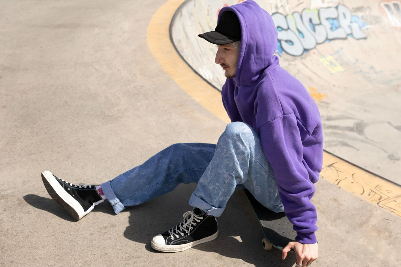 Skater wearing a purple streetwear hoodie, black sneakers, and a trucker hat, sitting on a skateboard at a skate park.
